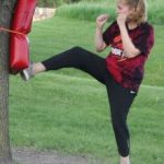 Woman in karate class kicking bag on tree outside in park.